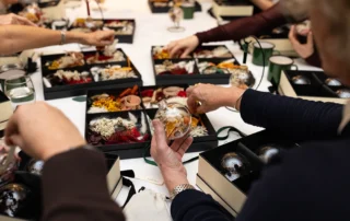 Close-up of hands filling a bauble with dried orange slices and flowers, over boxes of dried flower materials