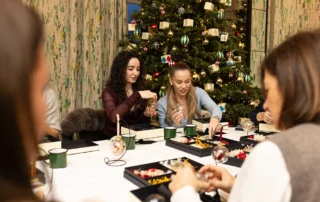 Two women filling their baubles with dried flowers at a Christmas masterclass, a decorated tree behind them